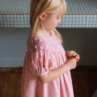 Pink cotton gingham girls’ Celeste Dress, hand‑smocked and embroidered, and puff sleeves from Elma.