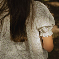Elma Delphi Blouse in Haru Blossom Iris, featuring delicate purple florals on white Japanese-milled cotton, a shirred neckline, puff sleeves with elasticated cuffs and picot lace trim.