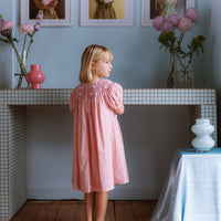 Pink cotton gingham girls’ Celeste Dress, hand‑smocked and embroidered, and puff sleeves from Elma.