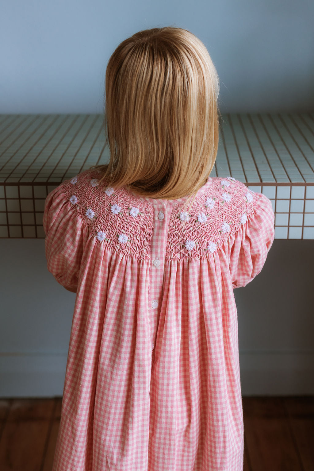 Pink cotton gingham girls’ Celeste Dress, hand‑smocked and embroidered, and puff sleeves from Elma.
