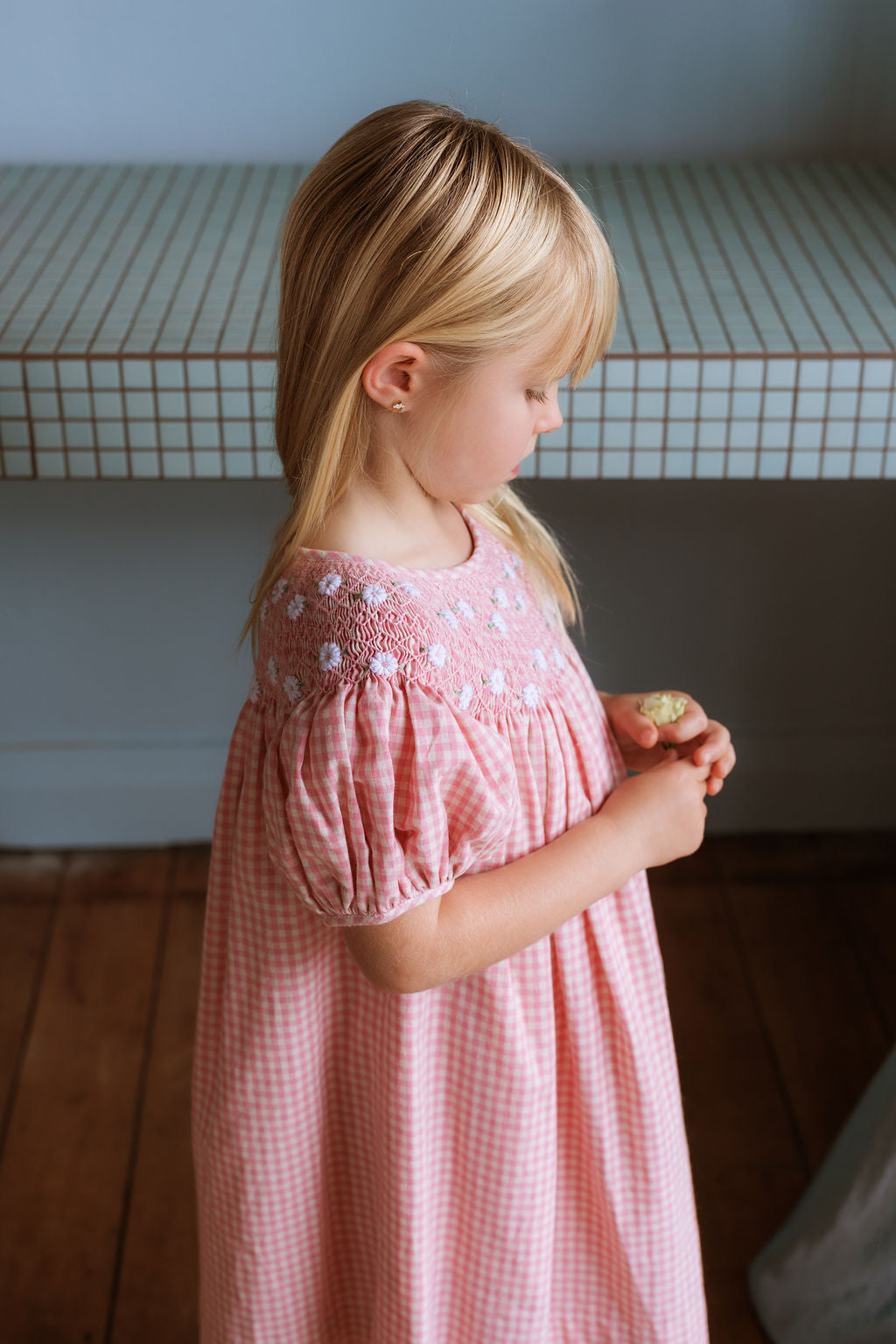 Pink cotton gingham girls’ Celeste Dress, hand‑smocked and embroidered, and puff sleeves from Elma.