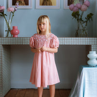 Pink cotton gingham girls’ Celeste Dress, hand‑smocked and embroidered, and puff sleeves from Elma.