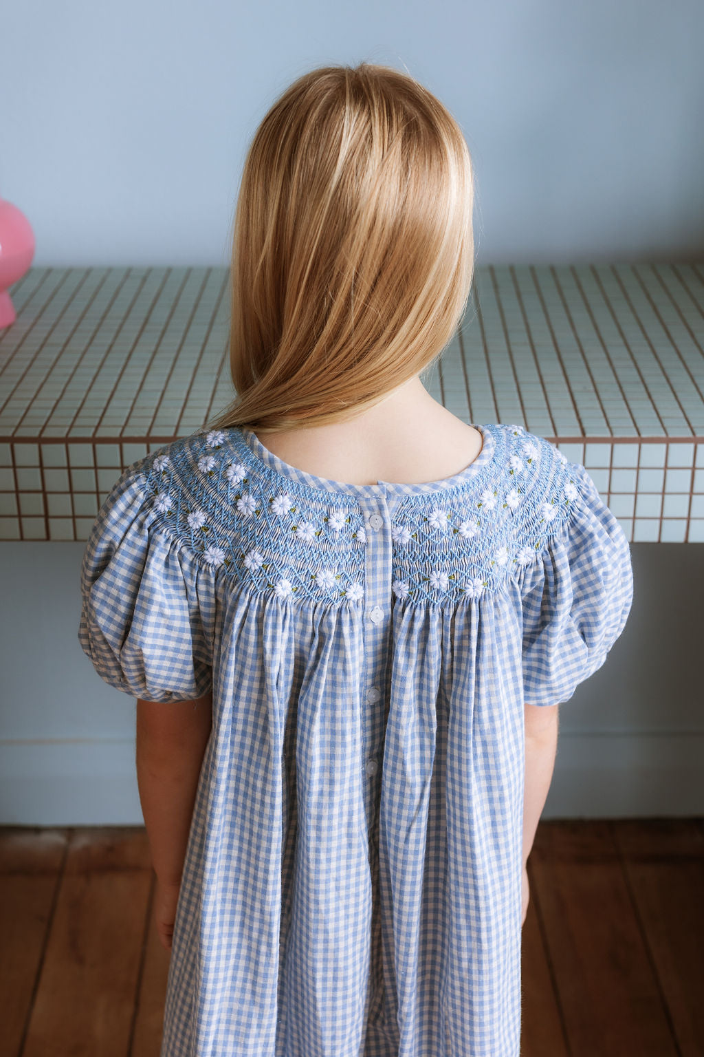 Celeste Dress in blue gingham, hand‑smocked and embroidered with petite daisies. Puff sleeves, button‑back and fully lined cotton.