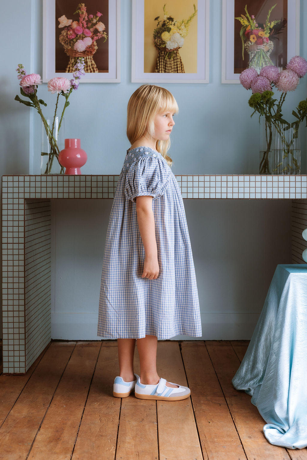 Celeste Dress in blue gingham, hand‑smocked and embroidered with petite daisies. Puff sleeves, button‑back and fully lined cotton.