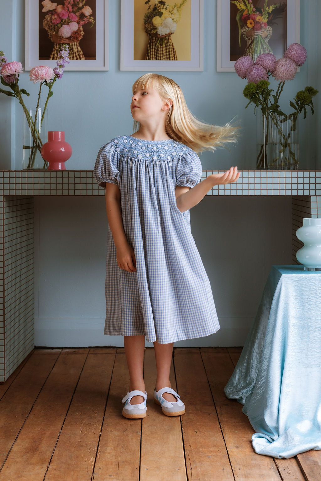 Celeste Dress in blue gingham, hand‑smocked and embroidered with petite daisies. Puff sleeves, button‑back and fully lined cotton.