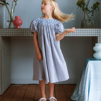 Celeste Dress in blue gingham, hand‑smocked and embroidered with petite daisies. Puff sleeves, button‑back and fully lined cotton.