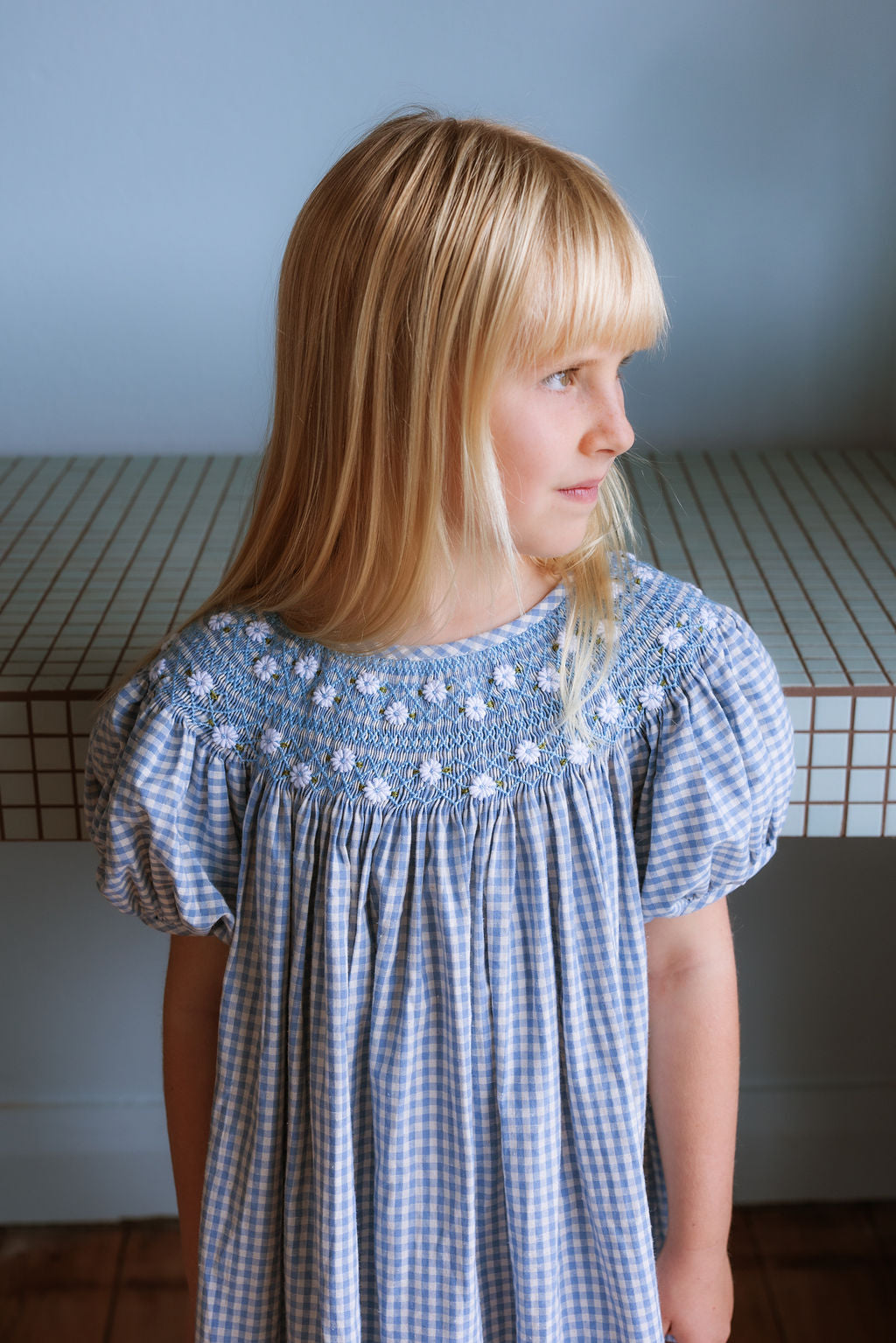 Celeste Dress in blue gingham, hand‑smocked and embroidered with petite daisies. Puff sleeves, button‑back and fully lined cotton.