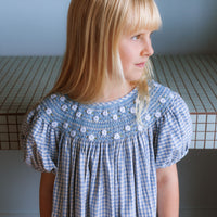 Celeste Dress in blue gingham, hand‑smocked and embroidered with petite daisies. Puff sleeves, button‑back and fully lined cotton.