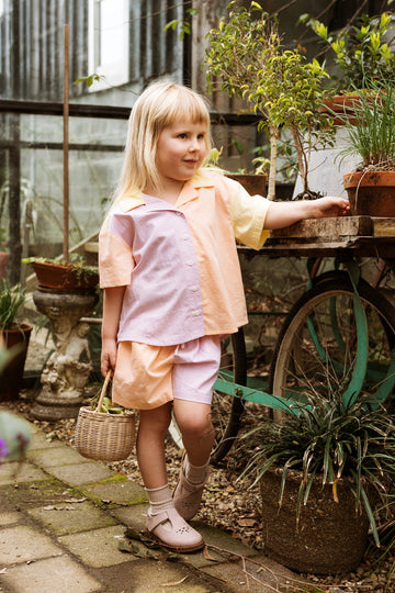 Girl standing in a courtyard surrounded by potplants. She is holding onto a wicker basket and wears a colourblocked cotton shirt and matching shorts.