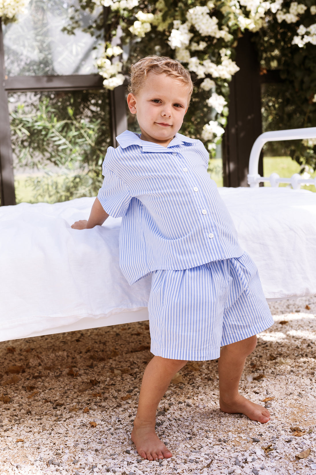 Little boy leaning against a bed wearing a blue stripe shirt and shorts set.