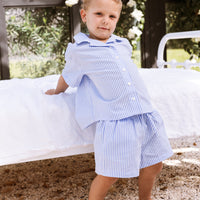 Little boy leaning against a bed wearing a blue stripe shirt and shorts set.