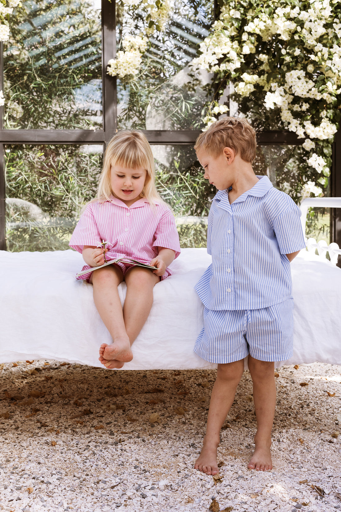 Girl and boy sitting on a bed reading a childrens story. One is wearing a pink stripe shirt and shorts set. The other is wearing a blue stripe shirt and shorts set.