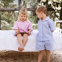 Girl and boy sitting on a bed reading a childrens story. One is wearing a pink stripe shirt and shorts set. The other is wearing a blue stripe shirt and shorts set.