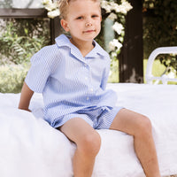 Little boy sitting on a bed wearing a blue stripe shirt and shorts set.