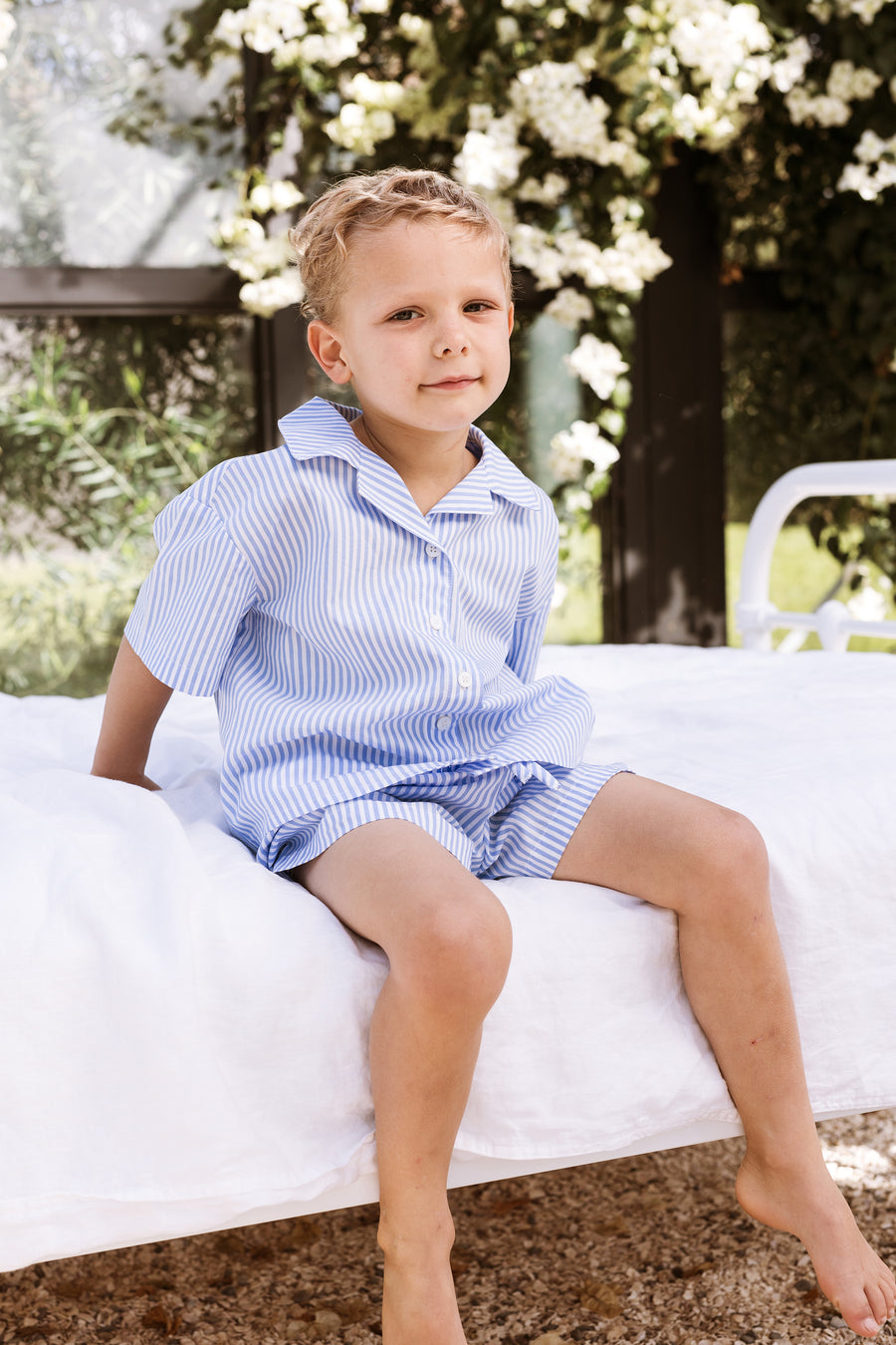 Little boy sitting on a bed wearing a blue stripe shirt and shorts set.