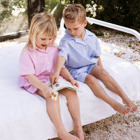 Girl and boy sitting on a bed reading a childrens story. One is wearing a pink stripe shirt and shorts set. The other is wearing a blue stripe shirt and shorts set.