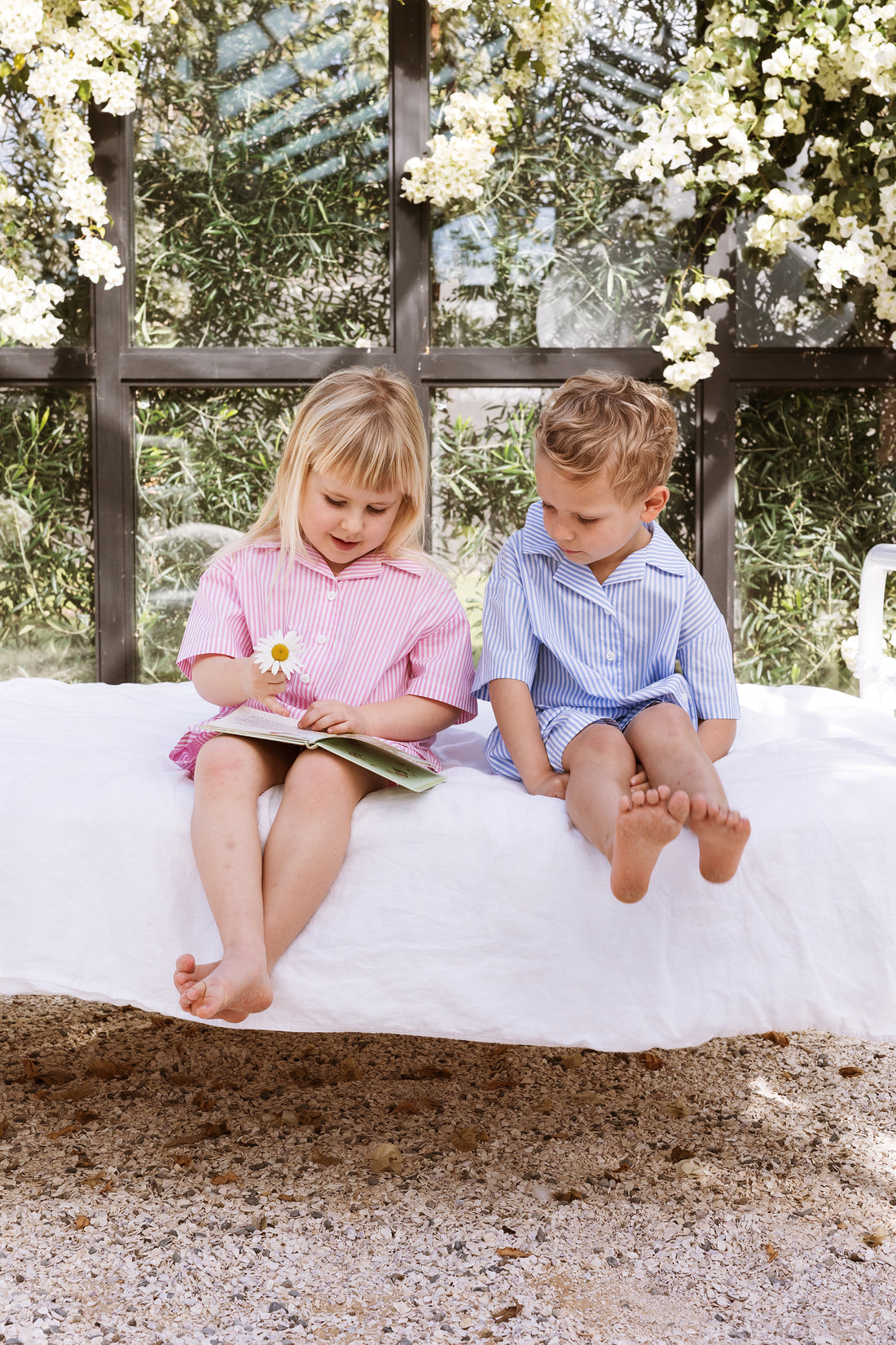 Girl and boy sitting on a bed reading a childrens story. One is wearing a pink stripe shirt and shorts set. The other is wearing a blue stripe shirt and shorts set.